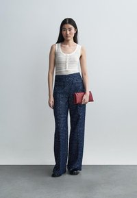 Naine seisab halli seina taustal, kandes valget varrukateta kootud topi, kõrge vöökohaga siniseid mustrilisi laia säärtega pükse, musti kingi ning hoiab punast käekotti.