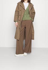 Brauner Trenchcoat, grüner Strickpullover, braune weite Hose und weiße Sneaker. Der Pullover hat Knöpfe und ein schichtartiges Design.