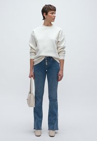 Balta džemperis, augsta vidukļa zilas zvana formas džinsi ar pogu detaļām, bēšas augstpapēžu kurpes un maza bēša somiņa.