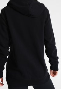 Czarna bluza z kapturem i długimi rękawami. Miękka, teksturowana tkanina, luźny krój oraz ściągacze na mankietach. Widoczne przeszycia na szwach z tyłu.