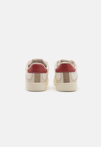 Adidași bej cu accente roșii și bej, textură din piele netedă și daim, formă rotundă, toc amortizat și talpă plată din cauciuc.