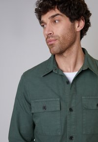 Grüne Baumwollbluse mit einem Kragen-Design, zwei Brusttaschen mit Knopfverschluss und einer strukturierten Oberfläche. Verstellbare Manschetten inklusive.