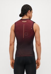 Sasniedzams bez piedurknēm sporta tops tumši sarkanā un melnā krāsā, ar gludu tekstūru, ģeometriskām izdrukām un zelta "VENUM" logo.