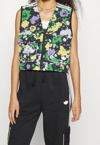 Blumenweste mit schwarzem Hintergrund, die grüne, gelbe und lila Blüten zeigt, mit Reißverschluss und zwei vorderen Taschen; darüber schwarze Jogginghosen getragen.