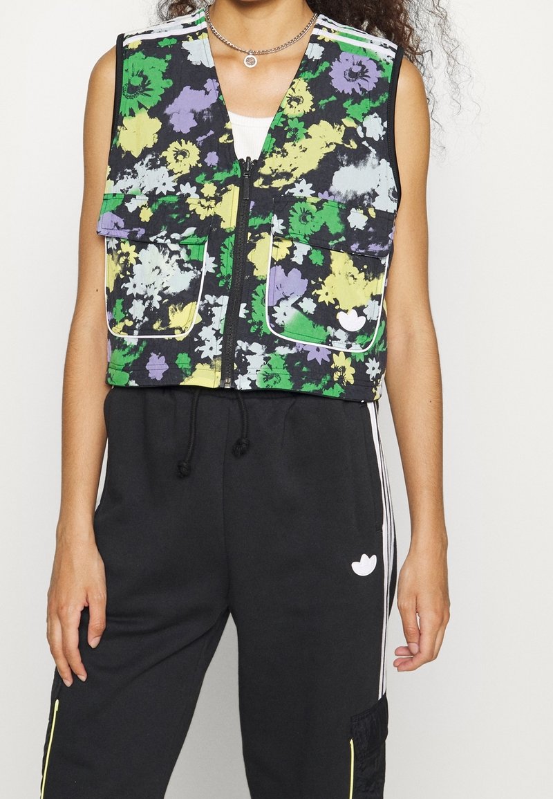 Blumenweste mit schwarzem Hintergrund, die grüne, gelbe und lila Blüten zeigt, mit Reißverschluss und zwei vorderen Taschen; darüber schwarze Jogginghosen getragen.