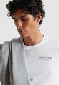 Witte katoenen t-shirt met zwart "FARAH" logo, gedragen onder een opgevouwen lichtgrijze sweatshirt, met een zachte textuur en een relaxte pasvorm.