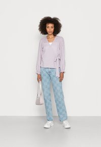Blusa envolvente cheque lilás e branca com mangas franzidas, combinada com jeans claros de xadrez azul e uma pequena mala rosa. Ténis brancos.