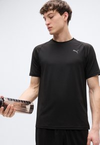 Μαύρο αθλητικό μπλουζάκι με κοντά μανίκια και κομψό σχέδιο. Διαθέτει ύφασμα που απομακρύνει την υγρασία και διακριτικό λογότυπο. Κρατά ένα διάφανο μπουκάλι νερού.