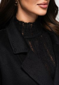 Manteau en laine noire avec un col à revers cranté et des coutures visibles ; porté sur un pull noir texturé à col haut avec des détails côtelés.