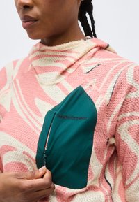 Persoană îmbrăcată într-un polar roz și crem cu model, având un buzunar la piept cu fermoar, de culoare teal, etichetat „Peak Performance”.