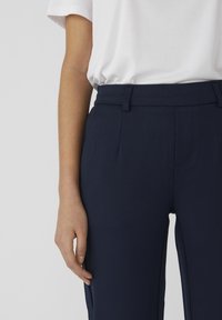 Pantalons ajustés marine en tissu lisse, présentant un devant plat, des poches latérales et des passants de ceinture, portés avec une chemise blanche unie.