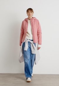 Roze gewatteerde jas met een capuchon, witte trui eronder, lichtblauwe wijde jeans en een gestreept shirt geknoopt om de taille, aangevuld met witte sneakers.