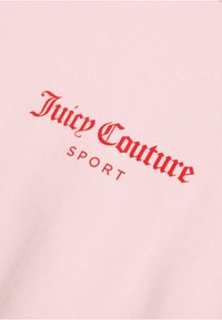 Rozā kokvilnas audums ar sarkaniem ekrāna drukas burtiem, kas lasa "Juicy Couture" virs vārda "SPORT" boldā tipografijā.