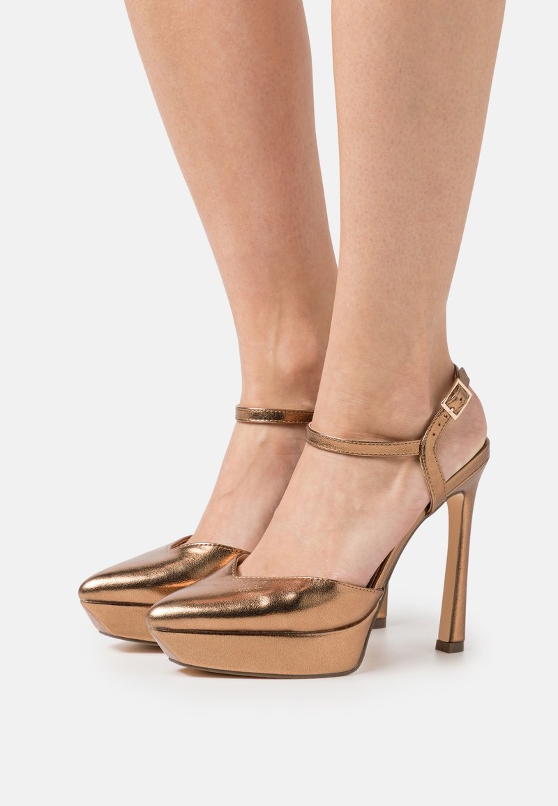 Pieds portant des escarpins métalliques bronze à bouts pointus avec brides à la cheville et semelles compensées sur un fond uni.