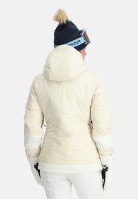 Weiße isolierte Jacke mit Kapuze, die ein gestepptes Design und einen kontrastierenden weißen Streifen an den Ärmeln aufweist. Dazu eine marineblaue Mütze mit Bommel.
