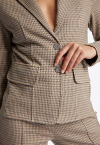 Blazer a motivi houndstooth marrone e grigio con due tasche frontali e bottoni decorativi. Tessuto strutturato con vestibilità su misura.