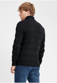 Maglione nero lavorato a maglia con colletto alto, caratterizzato da strisce orizzontali in texture. Abbinato a jeans in denim blu.