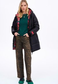 Čierna pérová bunda s kvetinovou podšívkou, zelený sveter a nohavice s leopardím vzorom. Oblečenie je doplnené zelenou obuvou, čím vytvára odvážny kontrast.