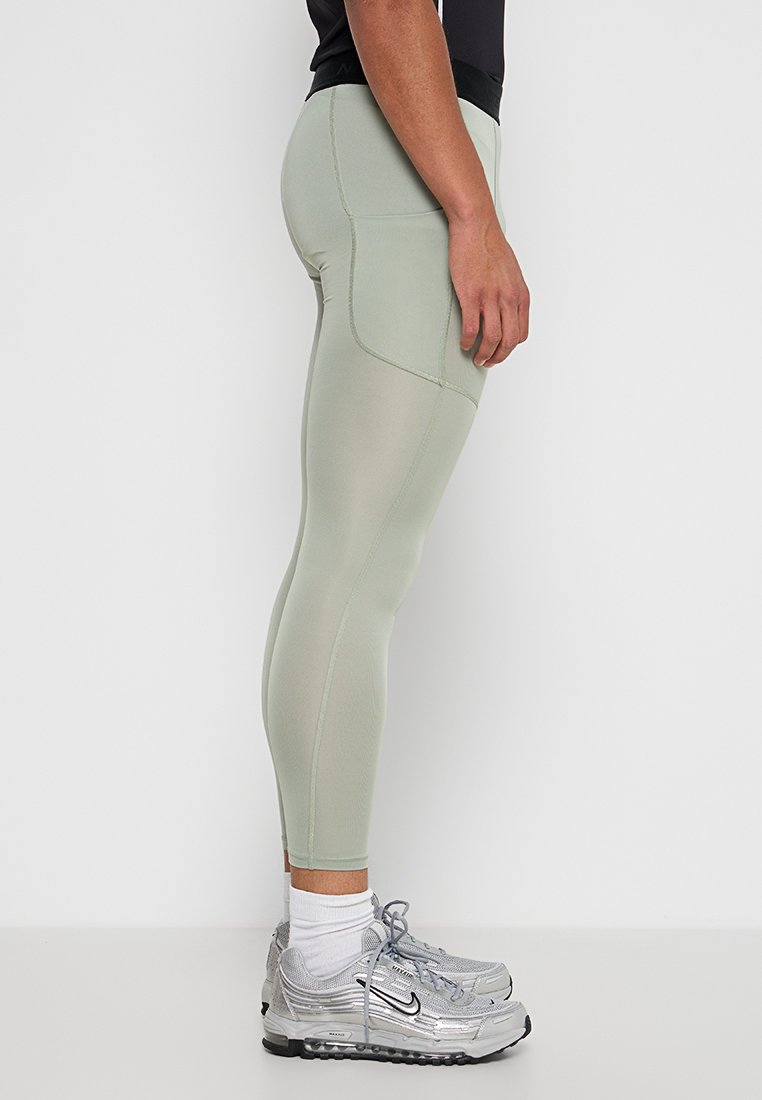 Leggings verde claro con una textura suave, que cuentan con un bolsillo lateral y un diseño ajustado. Combinados con zapatillas deportivas grises.