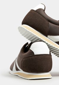 Scarpe da ginnastica marroni con una texture scamosciata, caratterizzate da accenti bianchi, un profilo basso e una suola bianca rigata. La vista posteriore evidenzia i materiali a contrasto.