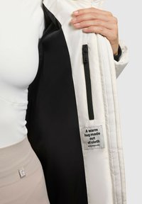 White quilted jacket with a black interior, featuring a side zipper and a fabric tag with a printed message. Smooth texture and clean design.