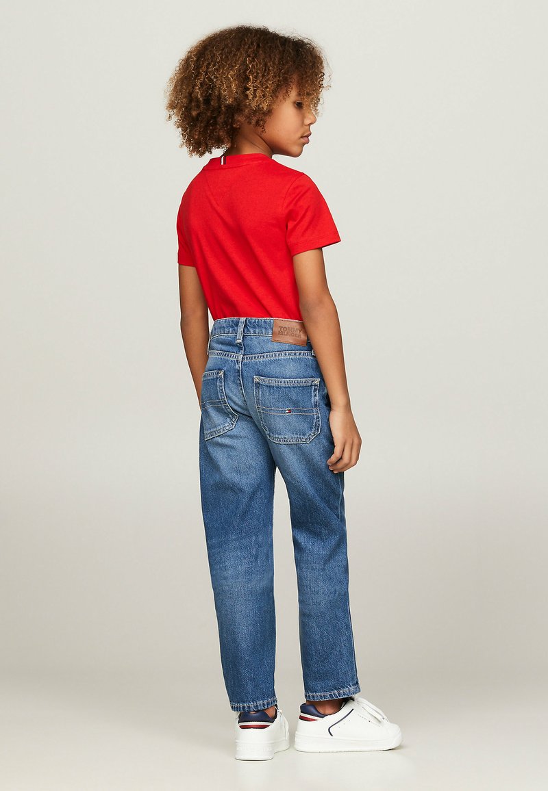 T-shirt rosso a maniche corte abbinato a jeans in denim blu. I jeans presentano un'alta vita, tasche posteriori e una toppa in pelle marrone del marchio sulla vita.