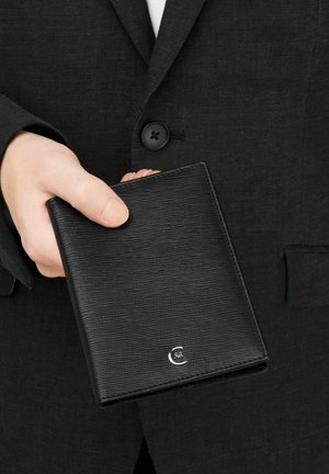 Ruka držící zavřenou černou texturovanou peněženku s malým stříbrným logem, v černém obleku s zapnutým knoflíkem.