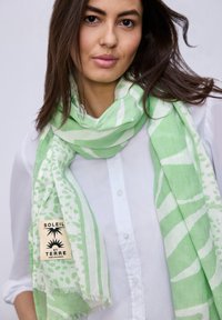 Femme portant une chemise blanche boutonnée et une écharpe à motifs verts et blancs portant l'inscription "SOLEIL ET TERRE" sur un fond uni.