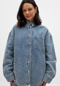 Blue denim shirt with a textured finish, button front, collar, and one chest pocket. Features loose fit and slightly frayed hems.