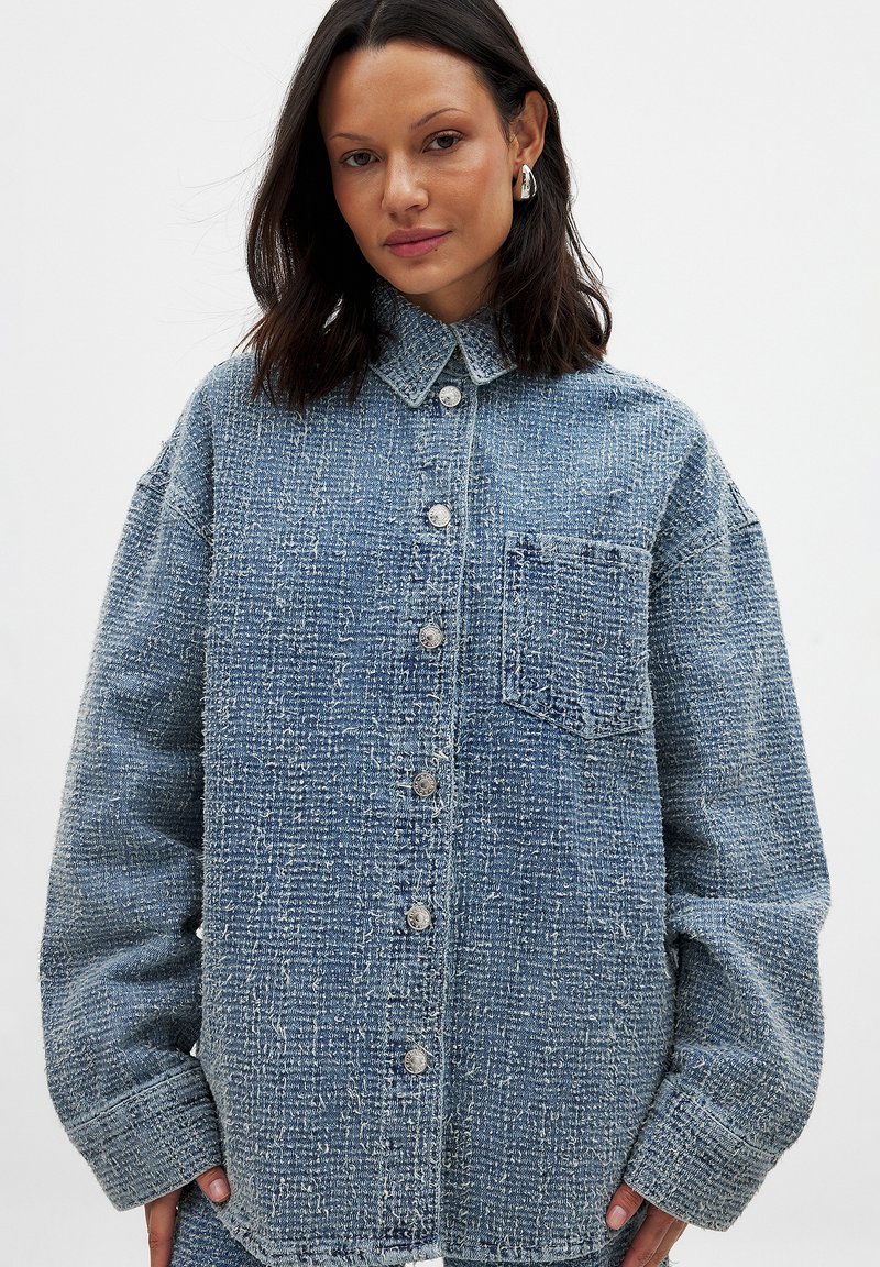 Blue denim shirt with a textured finish, button front, collar, and one chest pocket. Features loose fit and slightly frayed hems.