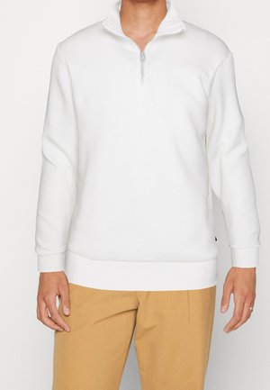 Personne portant un pull blanc à manches longues avec fermeture éclair au quart et un pantalon marron clair, debout devant un fond uni.