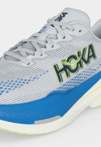 Λευκό και μπλε παπούτσι τρεξίματος Hoka με γκρι κορδόνια και φωτεινό κίτρινο και μαύρο λογότυπο στην πλαϊνή πλευρά.