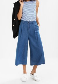 Denim culottes met een hoge taille en wijde benen, in een middelblauwe wassing. Gecombineerd met een gestreept mouwloos shirt en witte instap schoenen.