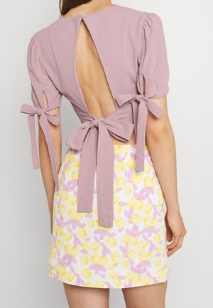 Donna che indossa un top lavanda con schiena scoperta e fiocchi legati sulle maniche e in vita, abbinato a una gonna con motivo floreale giallo e rosa.