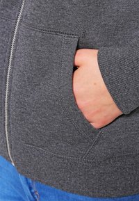 Felpa grigia con zip mostrata con una mano in una tasca anteriore. Il materiale è una stoffa testurizzata, con cuciture visibili e un polsino a coste.