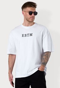 Weißes, übergroßes T-Shirt mit dem Aufdruck "ESTM" in Schwarz auf der Vorderseite, aus Baumwolle mit kurzen Ärmeln. Schwarze Hose und runde Sonnenbrillen runden den Look ab.