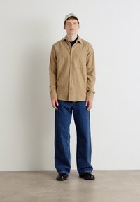 Cămașă butonată cu dungi maro deschis, pantaloni de blugi oversized mari, pantofi negri și o șapcă de culoare crem. Un outfit simplu și casual pe un fundal neutru.