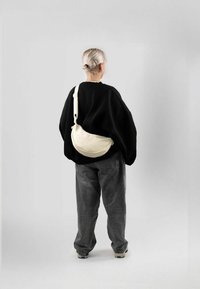 Hobo-Tasche aus cremefarbenem Stoff mit geschwungener Form und verstellbarem Tragegurt, getragen über einem schwarzen oversized Sweatshirt und grauen Hosen.