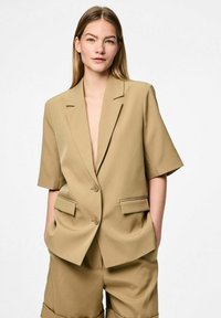 Blazer beige avec un décolleté en V profond, des manches courtes, deux poches avant et deux boutons. Texture de tissu lisse avec un design légèrement structuré.