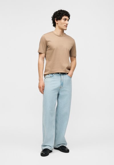 Homme aux cheveux bouclés debout, portant un t-shirt beige, un jean large bleu clair, des chaussures noires, une main dans la poche, regardant à droite.