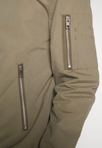 Olive green jacket sleeve with a textured fabric, featuring a zippered pocket and metallic zippers. Stitching visible on the seams.