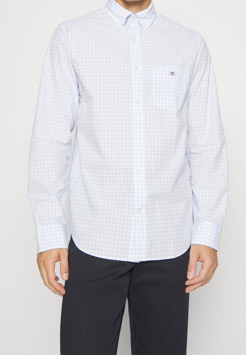Homme portant une chemise à carreaux bleu clair à manches longues et un pantalon foncé, debout devant un fond uni.