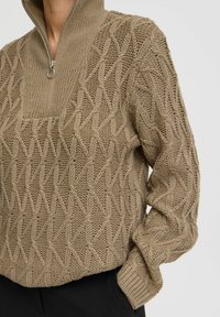 Beige gebreide pullover met een hoge kraag, voorzien van een halve rits en een gestructureerd ruitpatroon over de voorkant en de mouwen.
