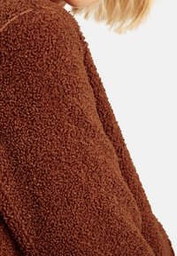 Brun, fluffig fleecejacka med en texturerad yta och en avslappnad passform. Tyget ser mjukt och mysigt ut, med en tjock, varm kvalitet.