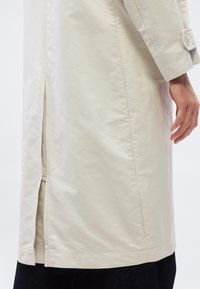 Μπεζ καμπαρντίνα με πλαϊνή σχισμή και μανσέτα με λεπτομέρεια κουμπιού, φορεμένη πάνω από μαύρο παντελόνι, εμφανίζοντας το χέρι ενός ατόμου που ακουμπά φυσικά δίπλα το υ.