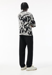 Person standing with arms crossed, wearing a black and white abstract patterned sweater, black pants, and white sneakers.