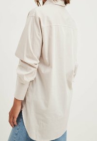 Light beige oversized shirt featuring vertical stripes, long sleeves, a button-down collar, and a curved hem. Smooth fabric texture.