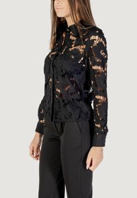 Zwarte kant shirt met bloemenpatroon, met lange mouwen en een knoopsluiting aan de voorkant. De stof is semi-doorzichtig, waardoor de onderliggende laag zichtbaar is.