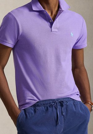 Personne portant un polo à manches courtes violet clair rentré dans un pantalon bleu à cordon, les mains dans les poches et une montre au poignet gauche.