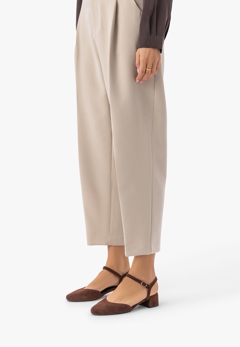 Naine, kes kannab beežika värvi kõrge vöökohaga laia säärega pükse ja pruune madala kontsaga pahkluuvööt kingi, seisab ühe käega lõdvestunult küljes.